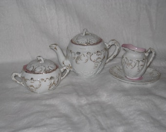 May include: A white porcelain tea set with gold filigree detailing and pink accents. The set includes a teapot, sugar bowl, creamer, and saucer. The tea set is on a white background.