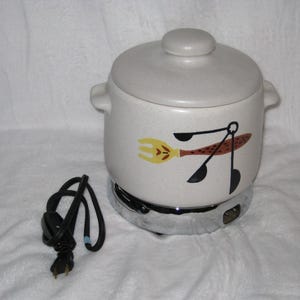 Può includere: Pentola elettrica a cottura lenta color crema con coperchio e manici. La pentola ha una base cromata e un cavo di alimentazione nero. Un disegno di forchetta, cucchiaio e spatola è sul lato. Il coperchio ha un piccolo pomello.