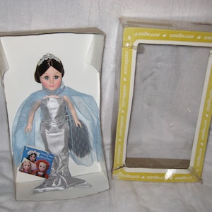 May include: A vintage doll in a silver mermaid gown, blue cape, and tiara, presented in its original box. The box is open, with the doll on the left and the empty box on the right. A small booklet with images of other dolls is included.