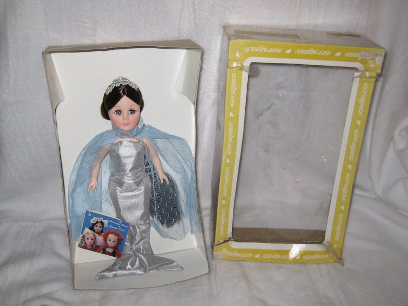 May include: A vintage doll in a silver mermaid gown, blue cape, and tiara, presented in its original box. The box is open, with the doll on the left and the empty box on the right. A small booklet with images of other dolls is included.