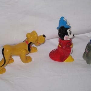 Puede incluir: Tres figuras de cer&aacute;mica: Pluto el perro amarillo, Mickey Mouse con una t&uacute;nica roja y sombrero azul, y un conejito gris con sombrero azul. Las figuras est&aacute;n sobre una superficie blanca.