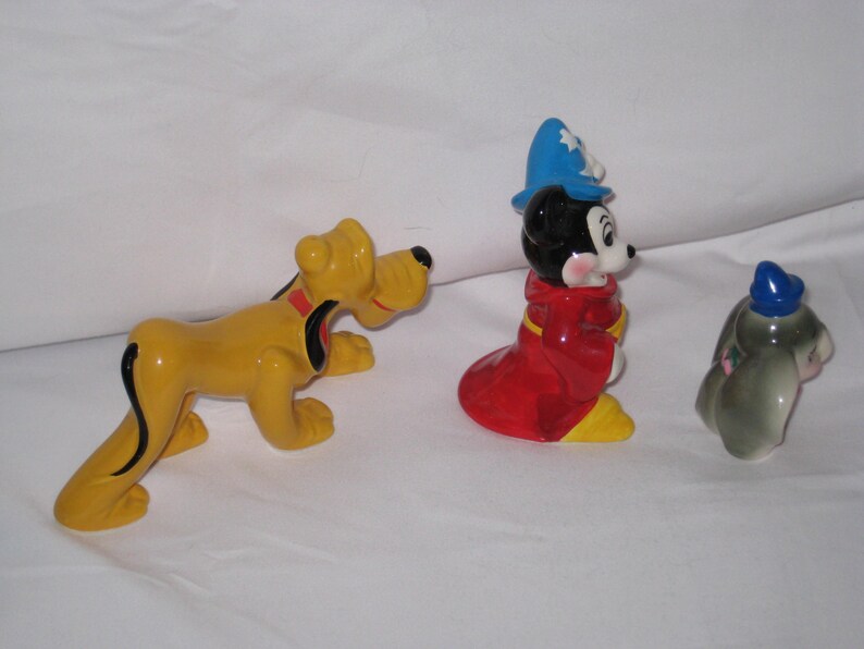Puede incluir: Tres figuras de cer&aacute;mica: Pluto el perro amarillo, Mickey Mouse con una t&uacute;nica roja y sombrero azul, y un conejito gris con sombrero azul. Las figuras est&aacute;n sobre una superficie blanca.