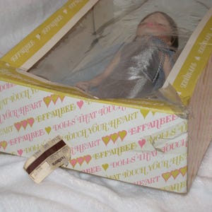 May include: A vintage Effanbee doll in its original box. The box is decorated with the text "DOLLS THAT TOUCH YOUR HEART" and a pattern of hearts. The doll is visible through a plastic window.