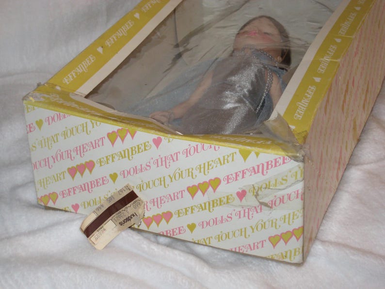 May include: A vintage Effanbee doll in its original box. The box is decorated with the text "DOLLS THAT TOUCH YOUR HEART" and a pattern of hearts. The doll is visible through a plastic window.