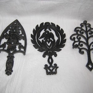 May include: Three black cast iron trivets with decorative designs. One is shaped like an iron, another features a bird and heart, and the third is a tree. These trivets are designed to protect surfaces from hot cookware.