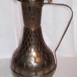 May include: A tall copper pitcher featuring a narrow neck, a wide rounded base, and a long curved handle. The exterior surface is decorated with a repeating honeycomb pattern of dark metallic ovals set against a polished copper background.