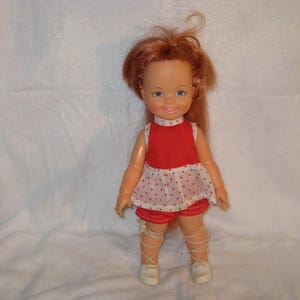 May include: A vintage doll with red hair, blue eyes, and a smiling expression. The doll wears a red and white polka-dot outfit with matching shorts and white shoes. The doll's legs are wrapped with white ribbons.
