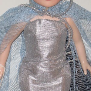 May include: A doll in a silver dress and a light blue mesh cape. A strand of pearls is draped around the neck. The doll's dress has a metallic sheen, and the cape is a delicate, airy fabric.
