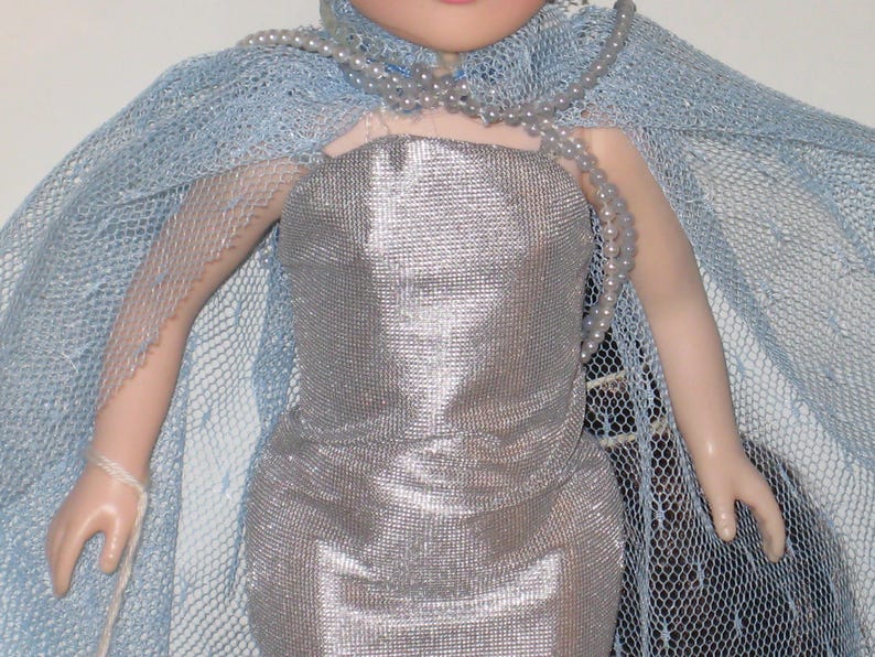 May include: A doll in a silver dress and a light blue mesh cape. A strand of pearls is draped around the neck. The doll's dress has a metallic sheen, and the cape is a delicate, airy fabric.