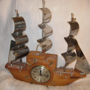 May include: A wooden ship-shaped clock with a round clock face and silver metal sails. The clock face has black numbers and hands. The ship's body is made of light brown wood.