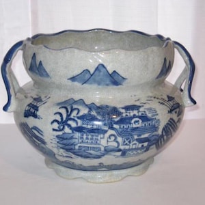 May include: A ceramic bowl with two handles, featuring a light grey crackle glaze finish and blue painted illustrations of traditional buildings, trees, and mountains. The bowl has a scalloped rim and a matching scalloped base design.