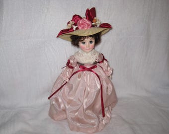May include: A vintage porcelain doll wearing a pink satin dress with a white lace collar and a straw hat adorned with pink and red flowers. The doll has blue eyes and brown curly hair, and is a collectible item.