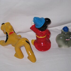 Puede incluir: Tres figuras de cer&aacute;mica: Pluto el perro amarillo, Mickey Mouse con abrigo rojo y gorro azul, y Dumbo el elefante gris con gorro azul. Las figuras est&aacute;n sobre una superficie blanca.