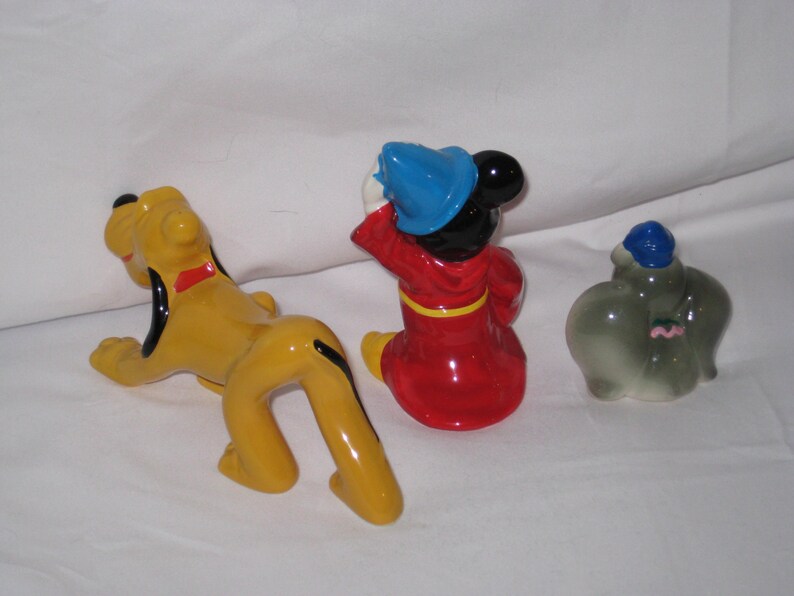 Puede incluir: Tres figuras de cer&aacute;mica: Pluto el perro amarillo, Mickey Mouse con abrigo rojo y gorro azul, y Dumbo el elefante gris con gorro azul. Las figuras est&aacute;n sobre una superficie blanca.