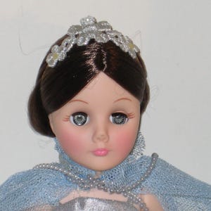 May include: A vintage doll with brown hair and a silver tiara. The doll has large, expressive eyes and rosy cheeks. She wears a blue lace shawl and a silver dress, with a pearl necklace. A classic collectible doll.