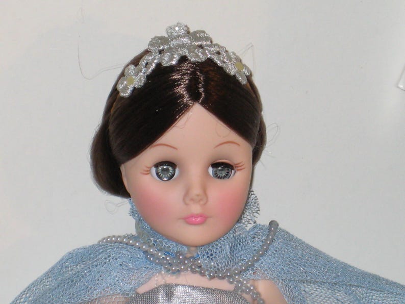 May include: A vintage doll with brown hair and a silver tiara. The doll has large, expressive eyes and rosy cheeks. She wears a blue lace shawl and a silver dress, with a pearl necklace. A classic collectible doll.