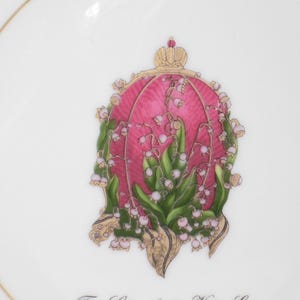 May include: White porcelain plate with a gold rim and a central design featuring a pink and gold floral emblem. The emblem includes green leaves, white flowers, and a gold crown.