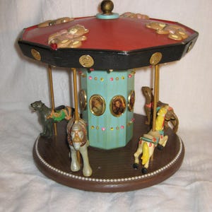 May include: A decorative carousel with a red and black roof, featuring small teddy bear figures. The carousel has a light blue center with oval portraits and is adorned with gold accents and colorful horses.