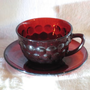 May include: A vintage ruby red glass teacup and saucer. The cup and saucer are a deep red colour with a textured, bubble-like pattern. The cup has a handle and sits on a matching saucer, creating a classic tea set.