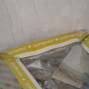 May include: A partially visible vintage Effanbee doll box. The box is yellow with the Effanbee logo and heart symbols. The box is open, revealing the doll inside, wrapped in plastic.