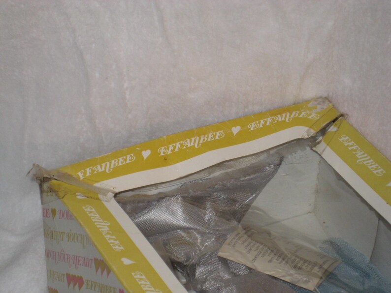 May include: A partially visible vintage Effanbee doll box. The box is yellow with the Effanbee logo and heart symbols. The box is open, revealing the doll inside, wrapped in plastic.
