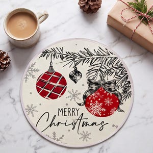 May include: A round placemat featuring a cream-colored background with a black ink illustration of pine branches, two red Christmas ornaments, and snowflakes. One ornament has a plaid pattern, the other a bow, with the text Merry Christmas.