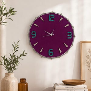 May include: A round wall clock with a deep purple velvet face, featuring teal-colored numbers at the 12, 3, 6, and 9 positions. The clock has silver-toned hands and is framed by a decorative silver rope border with small triangular markers.