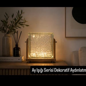 Op de afbeelding: Een vierkante glazen bloklamp met een getextureerd oppervlak, van binnenuit verlicht door warm licht. Het glazen blok wordt vastgehouden in een houten frame. De tekst "Ay Işığı Serisi Dekoratif Aydınlatma" staat in wit op een zwarte banner.