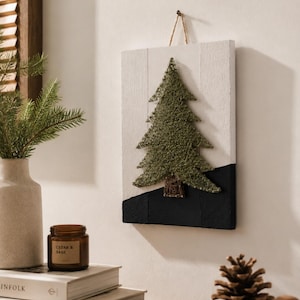 Op de afbeelding: Een rechthoekige wandhanger met een kerstboom van draadkunst in olijfgroen garen op een wit houten bord met een zwart geschilderde basis. De boom heeft een bruine draadkunststam en is opgehangen aan een dunne lus van jutekoord aan de bovenkant.