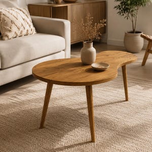 May include: A kidney-shaped wooden coffee table with a smooth, light brown finish and three tapered legs. The table surface holds a small, light-colored ceramic vase containing dried botanical stems and a small, shallow, matching ceramic bowl.