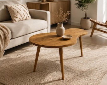 May include: A kidney-shaped wooden coffee table with a smooth, light brown finish and three tapered legs. The table surface holds a small, light-colored ceramic vase containing dried botanical stems and a small, shallow, matching ceramic bowl.