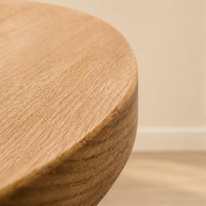 May include: Close-up of a wooden table corner. The table has a rounded edge and a light brown color. The wood grain is visible, adding texture to the surface. The table appears to be made of solid wood.
