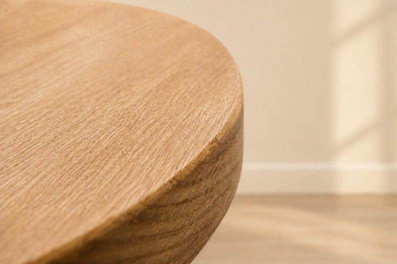 May include: Close-up of a wooden table corner. The table has a rounded edge and a light brown color. The wood grain is visible, adding texture to the surface. The table appears to be made of solid wood.