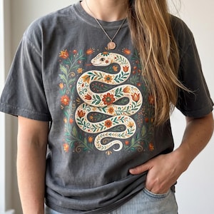 Könnte beinhalten: Ein anthrazitfarbenes Kurzarm-T-Shirt mit einer zentralen Grafik einer weißen Schlange, die in S-Form gewunden ist und mit orangefarbenen, roten und grünen Blumen- und Blattmustern verziert ist. Die Schlange ist von kleinen orangefarbenen und roten Blüten umgeben.