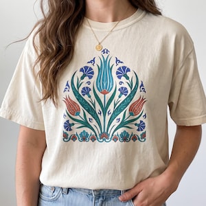 May include: A cream-colored short-sleeve t-shirt featuring a central graphic print of stylized tulips and carnations in shades of blue, teal, and terracotta. The floral design is symmetrical, with intricate leaf patterns and decorative borders.
