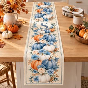 May include: A rectangular table runner featuring a repeating pattern of orange, blue, and white pumpkins interspersed with blue and cream flowers and green foliage. The center of the runner displays a large, dark blue capital letter S in a serif font.
