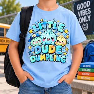 May include: A light blue t-shirt featuring a graphic of three cartoon dumplings with faces, surrounded by yellow stars and blue lightning bolts. The text reads "LITTLE DUDE DUMPLING" in a bubbly, colourful font. The design is centred on the chest.