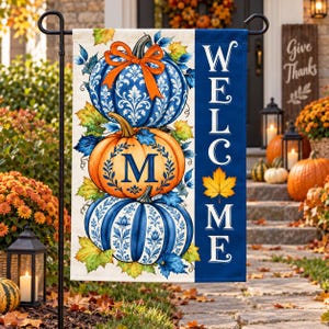 May include: A rectangular garden flag featuring a stack of three pumpkins decorated with blue and white floral patterns. The middle pumpkin displays the letter M. An orange bow sits atop the stack. A blue vertical stripe reads WELCOME.