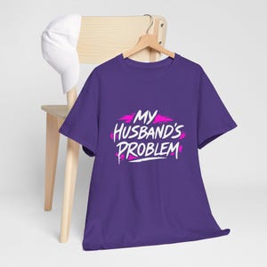 May include: A purple t-shirt with the phrase "MY HUSBAND'S PROBLEM" in white script with pink accents. The shirt is hanging on a wooden hanger. A white baseball cap rests on a wooden chair in the background.