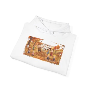 May include: A white hooded sweatshirt featuring a rectangular graphic print of the Peanuts characters playing in a pile of autumn leaves. The illustration includes Charlie Brown, Snoopy, and friends surrounded by orange trees and pumpkins.