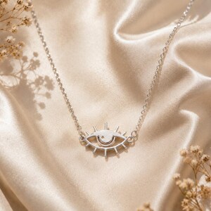 May include: A silver-toned necklace featuring a central pendant shaped like an eye with radiating lines resembling eyelashes. The pendant is attached to a delicate silver-toned chain, creating a cohesive piece of jewelry with a metallic finish.