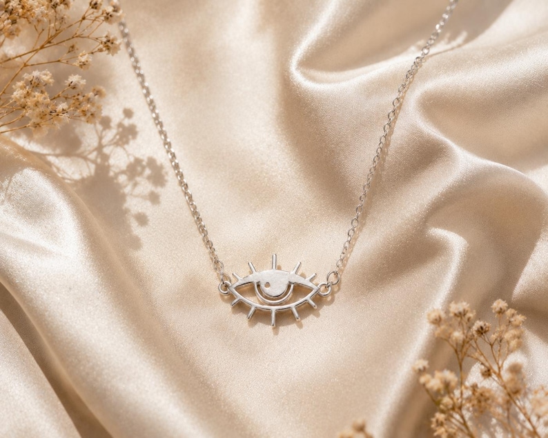 May include: A silver-toned necklace featuring a central pendant shaped like an eye with radiating lines resembling eyelashes. The pendant is attached to a delicate silver-toned chain, creating a cohesive piece of jewelry with a metallic finish.