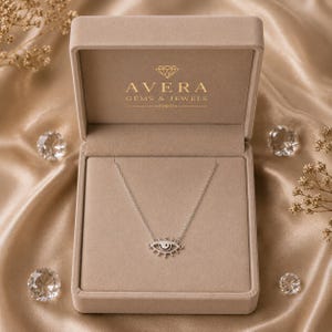 May include: A silver-toned necklace featuring an eye-shaped pendant with a central black dot, displayed inside a beige velvet jewelry box. The interior lid of the box is printed with the gold text Avera Gems & Jewels and a diamond icon.