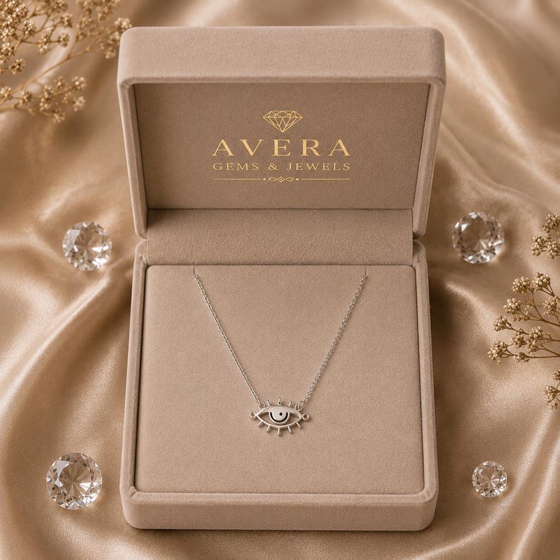 May include: A silver-toned necklace featuring an eye-shaped pendant with a central black dot, displayed inside a beige velvet jewelry box. The interior lid of the box is printed with the gold text Avera Gems & Jewels and a diamond icon.