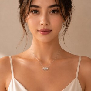 May include: A silver-toned necklace featuring a central pendant shaped like an eye with radiating lines resembling eyelashes. The pendant is suspended from a delicate, thin silver-toned chain that rests against the collarbone area of the wearer.