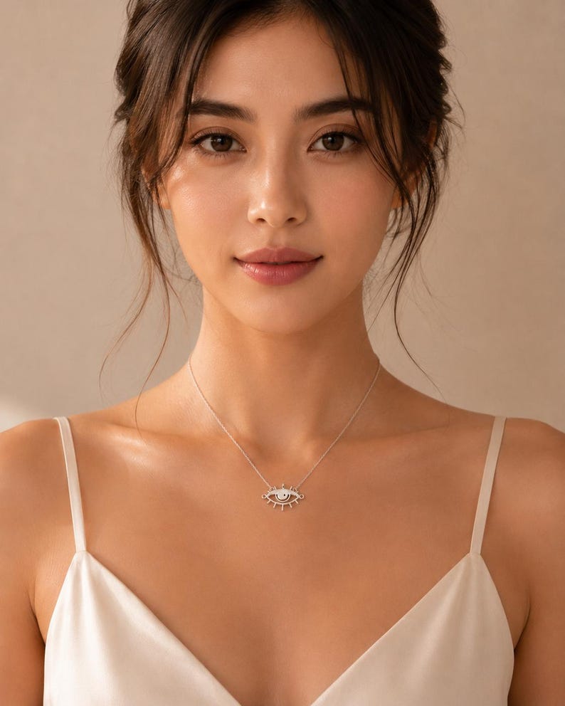 May include: A silver-toned necklace featuring a central pendant shaped like an eye with radiating lines resembling eyelashes. The pendant is suspended from a delicate, thin silver-toned chain that rests against the collarbone area of the wearer.