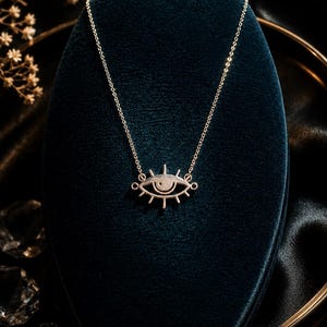 May include: A silver-toned necklace featuring a stylized eye pendant with radiating lashes. The pendant is suspended from a delicate, thin silver-toned chain. The design is symmetrical, with a central pupil and a clean, polished metallic finish.