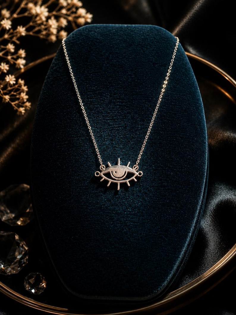 May include: A silver-toned necklace featuring a stylized eye pendant with radiating lashes. The pendant is suspended from a delicate, thin silver-toned chain. The design is symmetrical, with a central pupil and a clean, polished metallic finish.