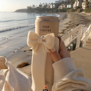 May include: A can of vanilla iced coffee with the text "Vanilla Iced Coffee Oatmilk" is held in a cream-coloured bow-adorned holder. The background shows a beach and ocean scene.