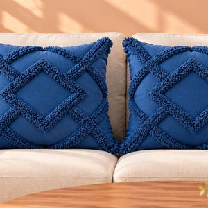 Puede incluir: Dos cojines cuadrados de color azul real intenso. Cada cojín presenta un patrón geométrico texturizado en relieve que consiste en formas de diamante y líneas interconectadas, creadas con detalles de tela afelpada y difusa en la parte frontal.